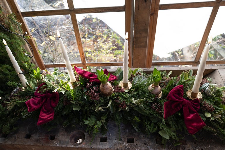 Dekorativt arrangemang med ljus, grangrenar, band och växter framför ett fönster.