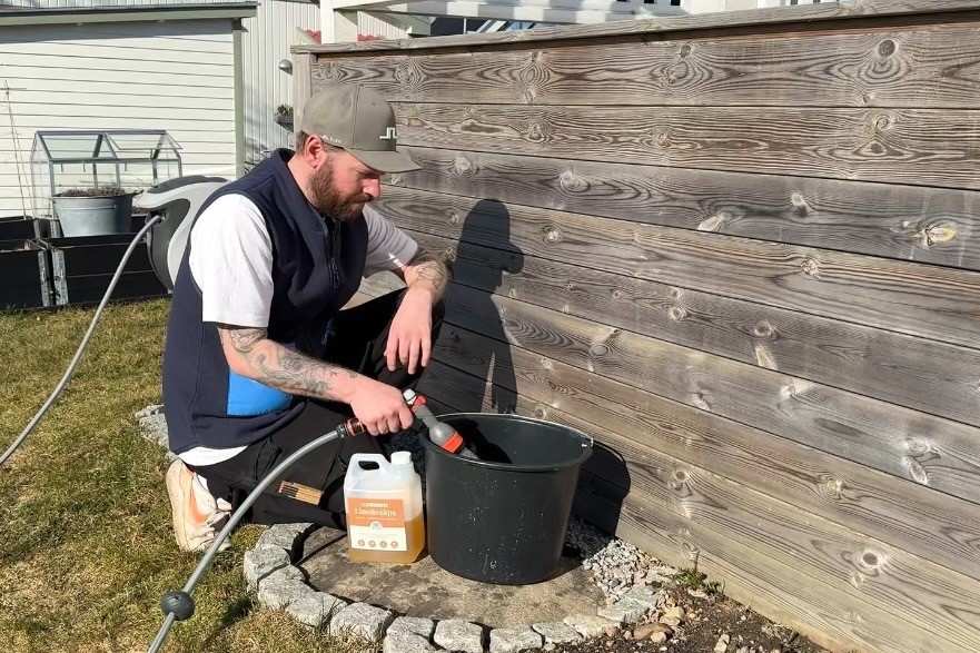 En man rengör ett trästaket i trädgården med trärengöring och fyller en hink med vatten med en trädgårdsslang.