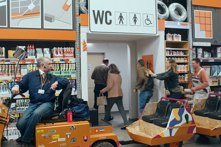Scen i en Hornbach-byggmarknad med kunder och en anställd på ett orange elfordon framför en toalettskylt