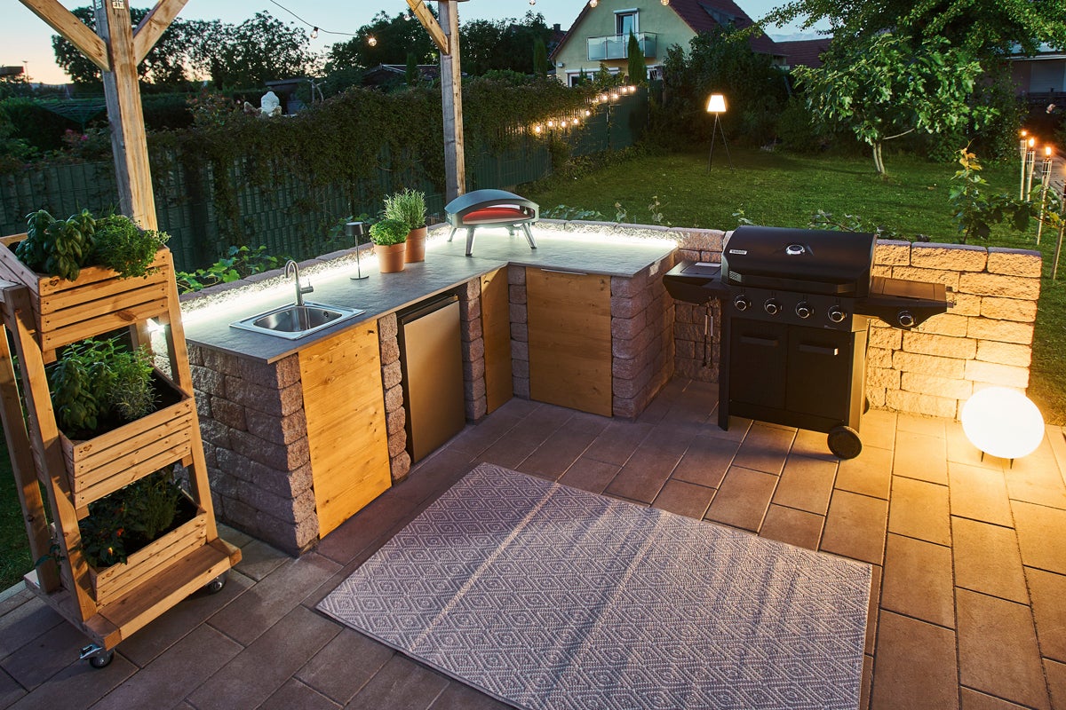 Utomhuskök med grill, diskho, pizzaugn och örtagård under en pergola på en terrass