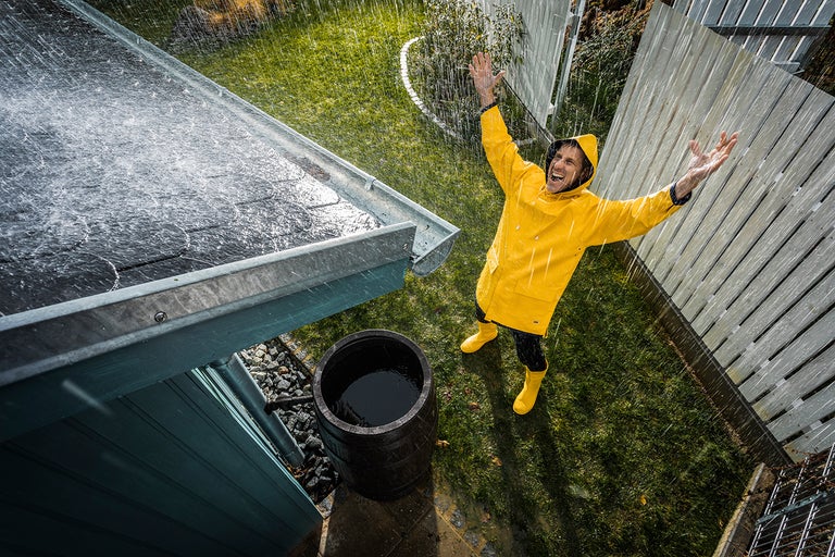 Regnvattenuppsamlingsfat bredvid ett tak med hängränna och man med regnkläder i trädgården