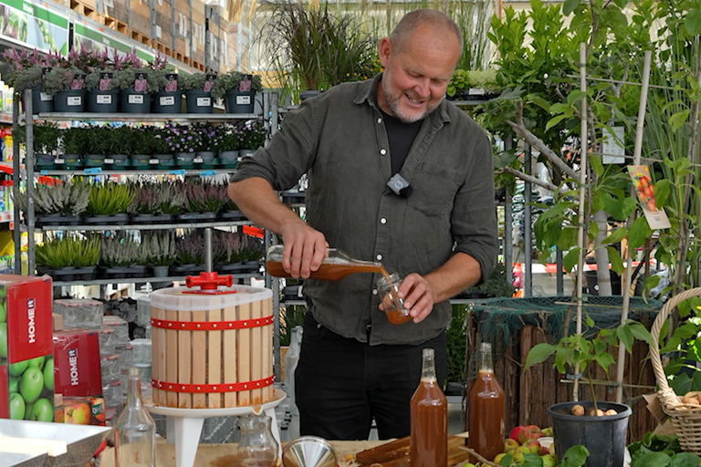 En man häller nypressad äppeljuice från en glasflaska i ett glas bredvid en fruktpress i trä i ett trädgårdscenter.
