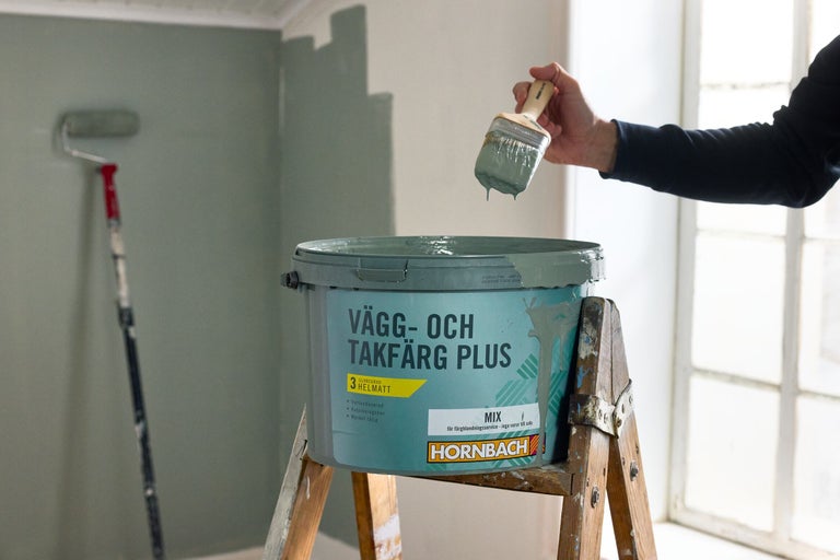 En person målar en vägg med Hornbach vägg- och takfärg Plus. Öppen färgburk på en trästege med en pensel och en färgroller mot väggen.