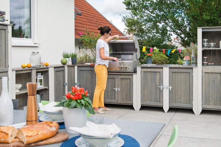 Utomhuskök med kvinna vid grillen, bord med porslin och mat i förgrunden