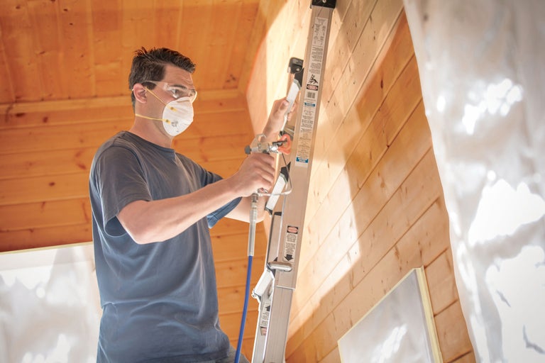 Man med skyddsmask och skyddsglasögon på en stege som sprutar en trävägg