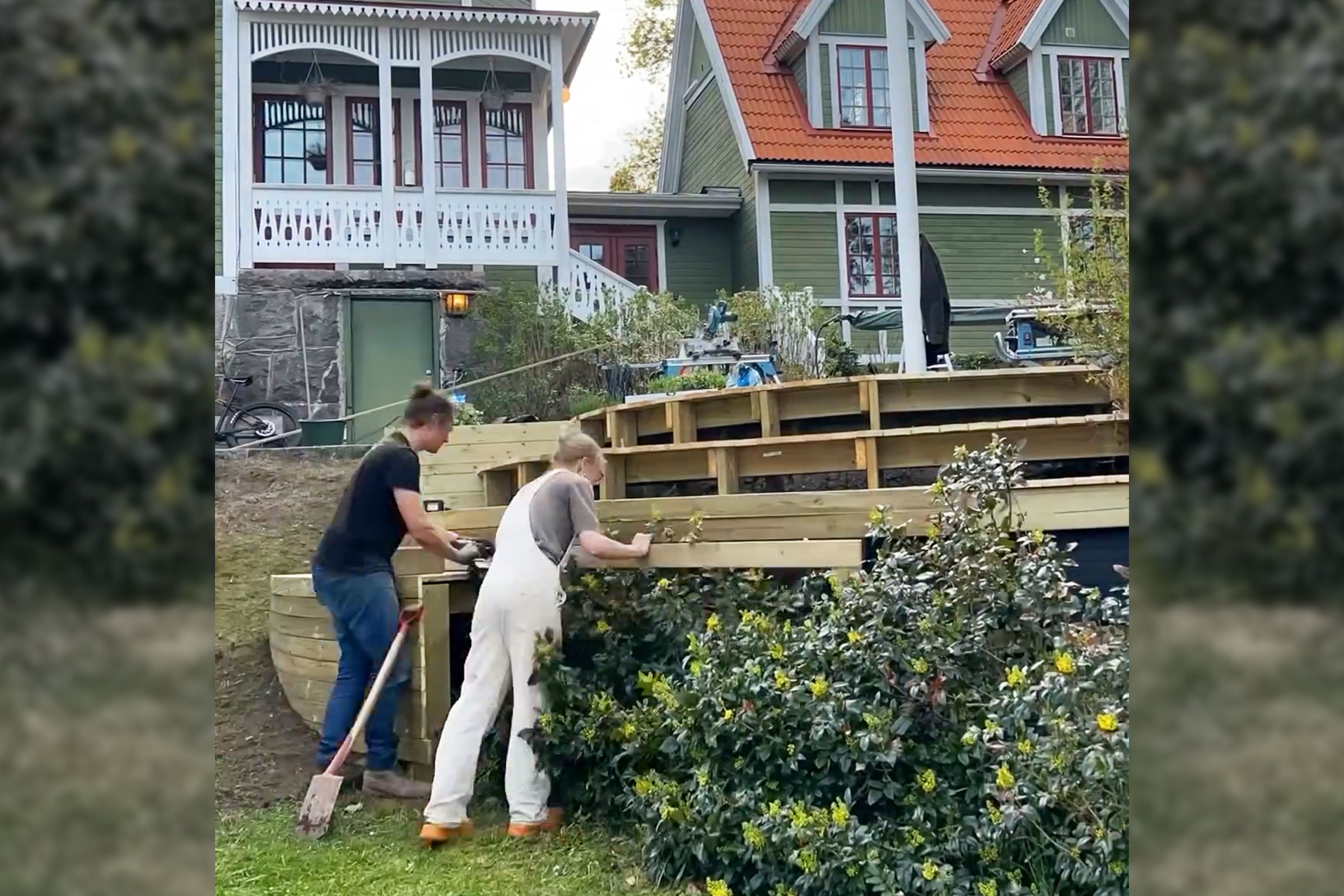 Två personer bygger en träterrass i en trädgård med bostadshus