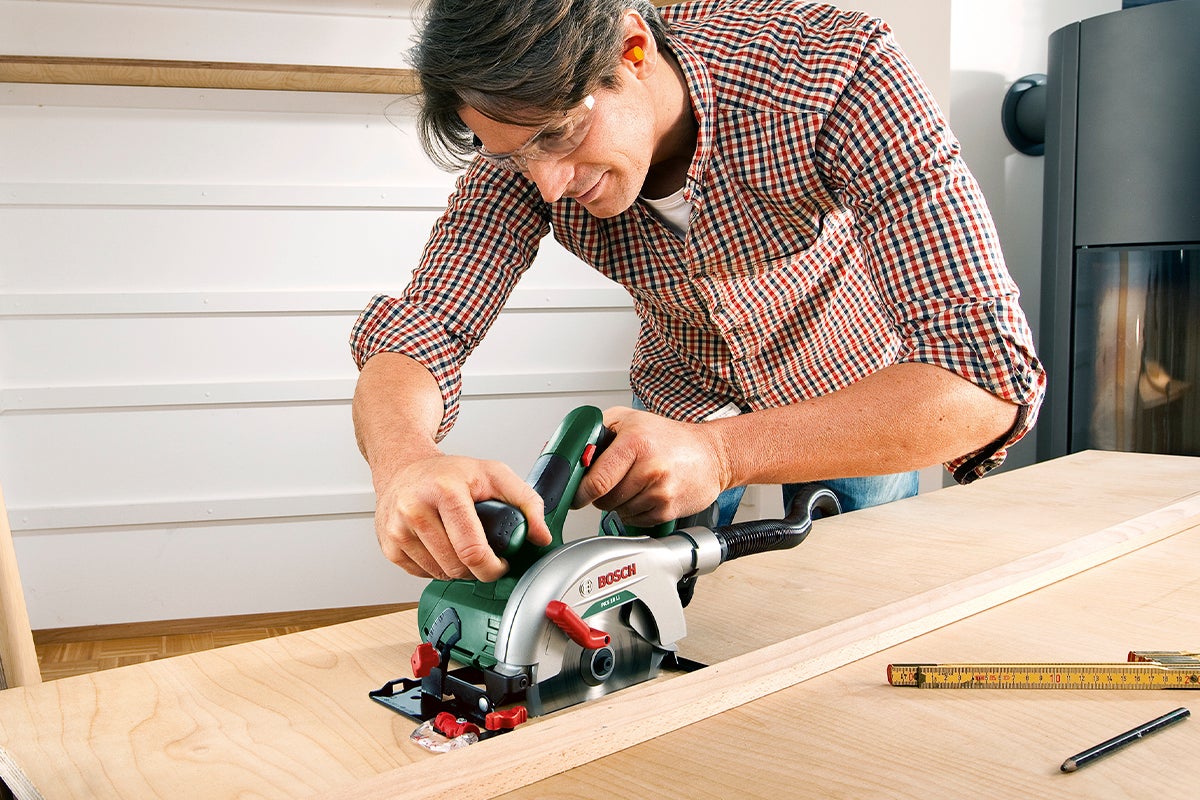 Man sågar träbräda med en Bosch batteridriven cirkelsåg. En tumstock och en penna ligger på bordet.