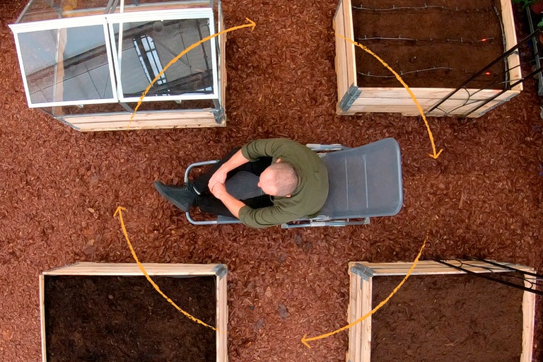 Ovanifrån ser man en man på en solstol mellan upphöjda rabatter och ett växthus