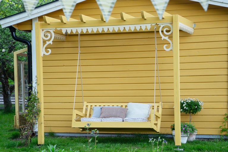 Trädgårdsscen med en gul träsving, dekorerad med kuddar, framför ett gult trähus med vit dekor och vimpel.