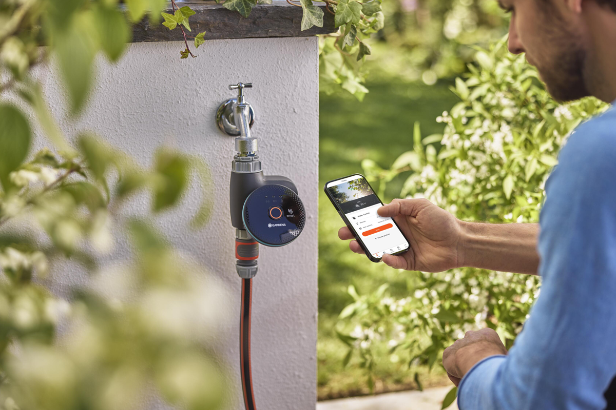 Man styr en smart bevattningsdator från Gardena på en utomhuskran med en app i sin smartphone i trädgården.