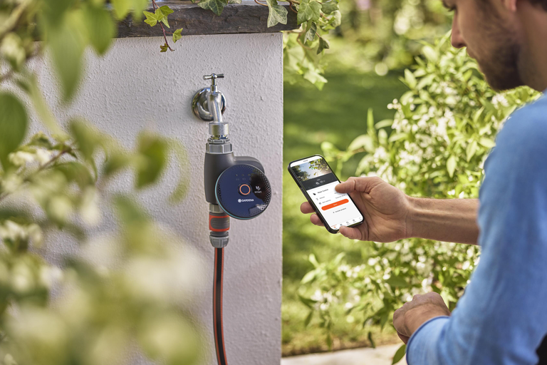 Man styr en smart bevattningsdator från Gardena på en utomhuskran med en app i sin smartphone i trädgården.