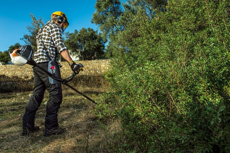 En man med skyddskläder och en röjsåg trimmar en häck i trädgården.