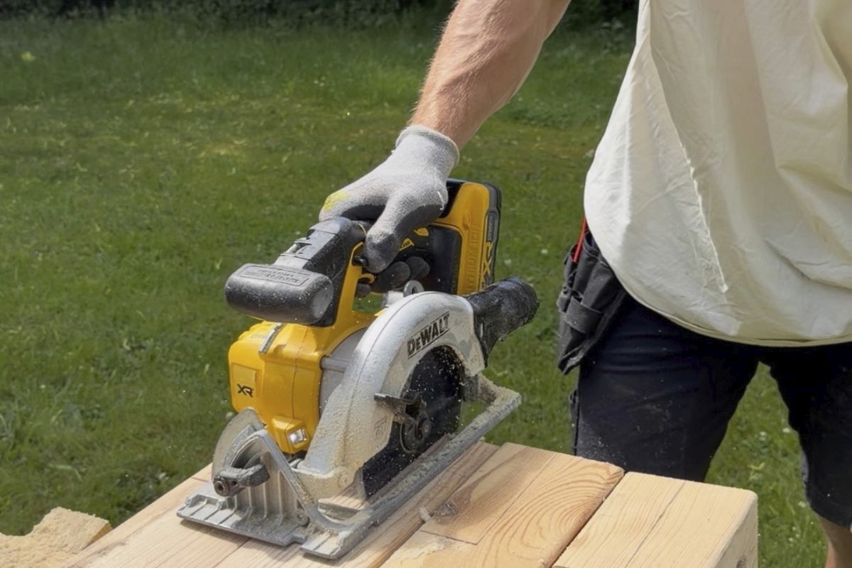 En person använder en sladdlös DeWalt cirkelsåg för att såga träplankor utomhus.