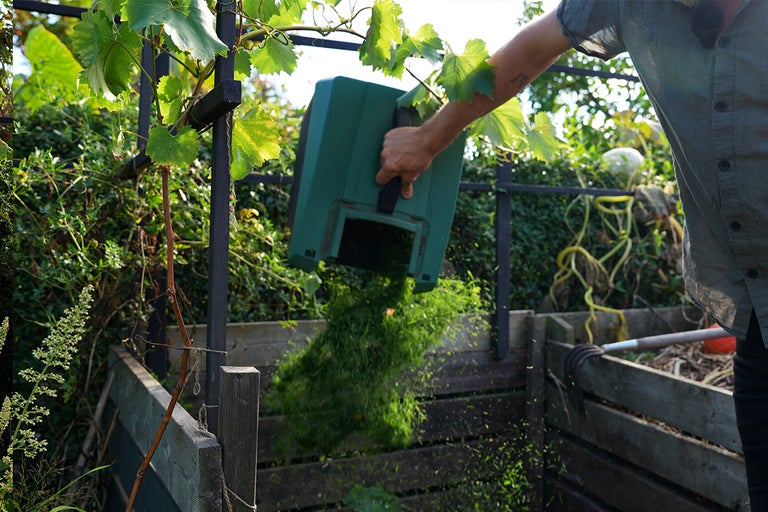 Person tömmer grönt avfall från en kompostbehållare i en öppen träkompost.