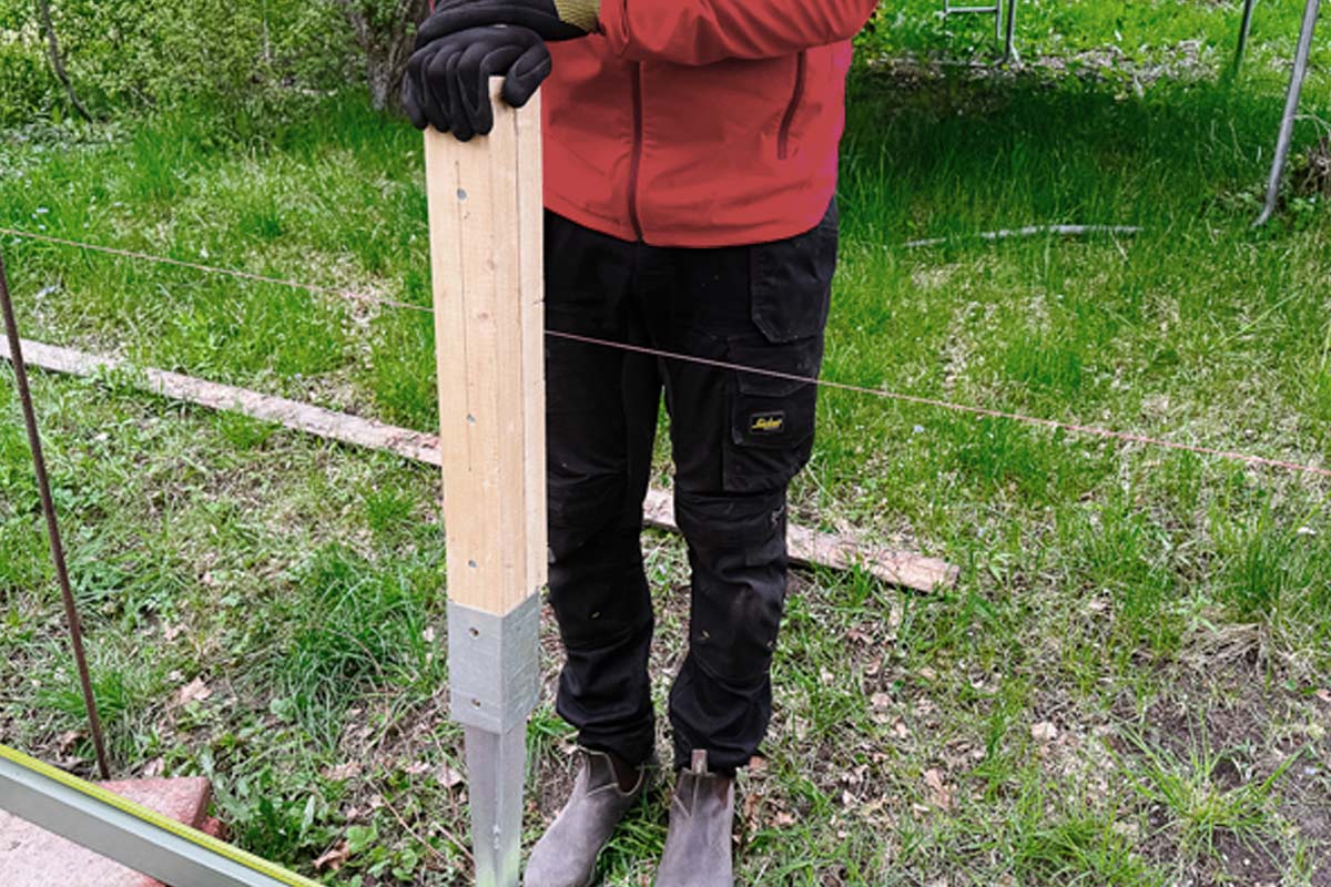 Person håller trästolpe med nedslagningshylsa i trädgården.