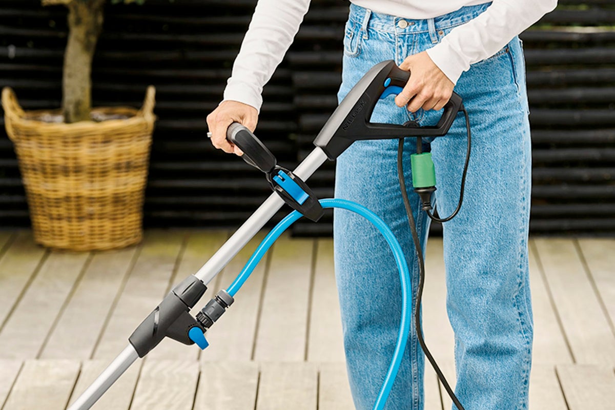 En person använder en Nilfisk terrasstvätt på en ljus träaltan. Maskin med svart handtag, Nilfisk Logo och blå slang.