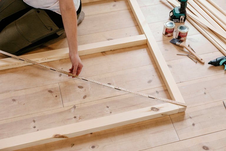 Person mäter träram med tumstock, med verktyg, träribbor och träskyddsfärg på trägolv