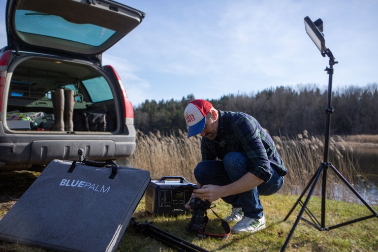 En man förbereder fotoutrustning utomhus, inklusive en kraftstation, en kameraväska och ett stativ.
