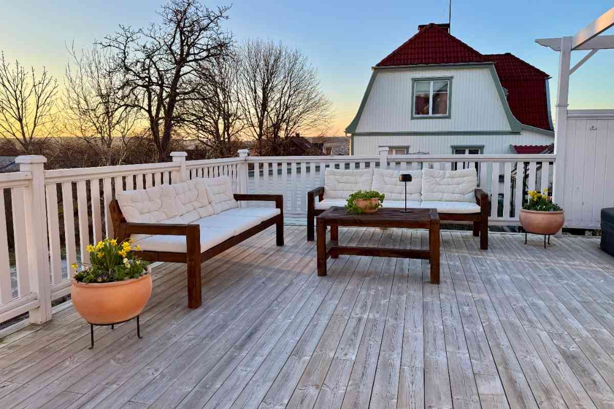Ljus träterrass med mörka trädgårdssoffor, vita dynor, träbord, vitt räcke och blomkrukor i terrakotta framför ett bostadshus.