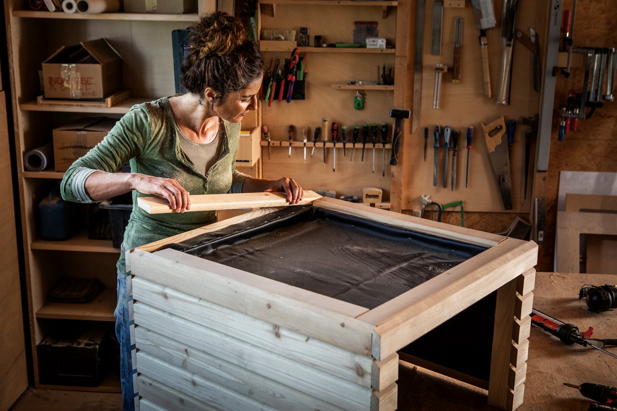 En kvinna bygger en odlingslåda av ljust trä med svart innerduk i en verkstad. Verktyg och hyllor syns i bakgrunden.