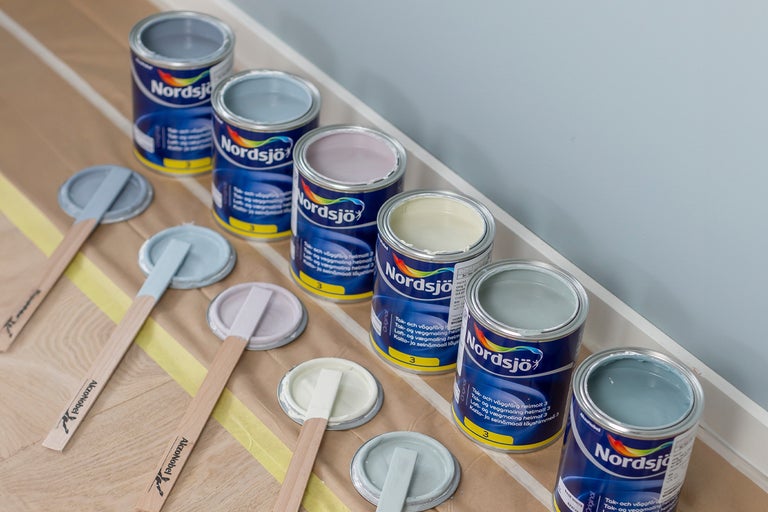 Färgval: Flera färgburkar av märket Nordsjö med färgprover och träpinnar för att välja rätt färg.