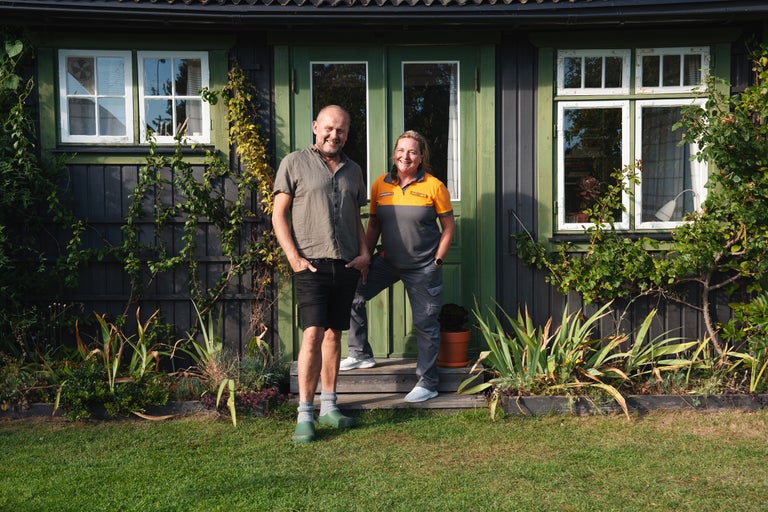 Två personer står framför ett hus med träväggar och fönster med vita ramar. Huset är omgivet av vegetation.