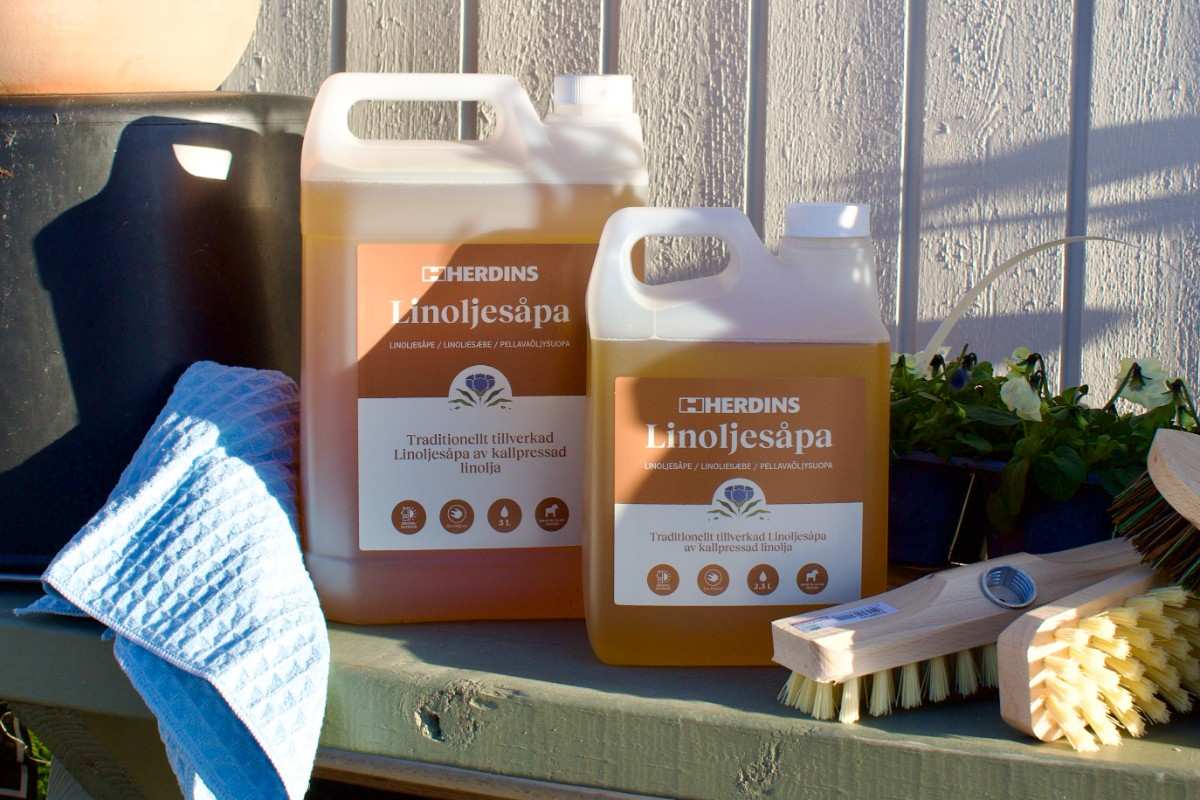 Två dunkar Herdins Linoljesåpa med en blå torkduk och skurborstar av trä på en träyta i solljus.