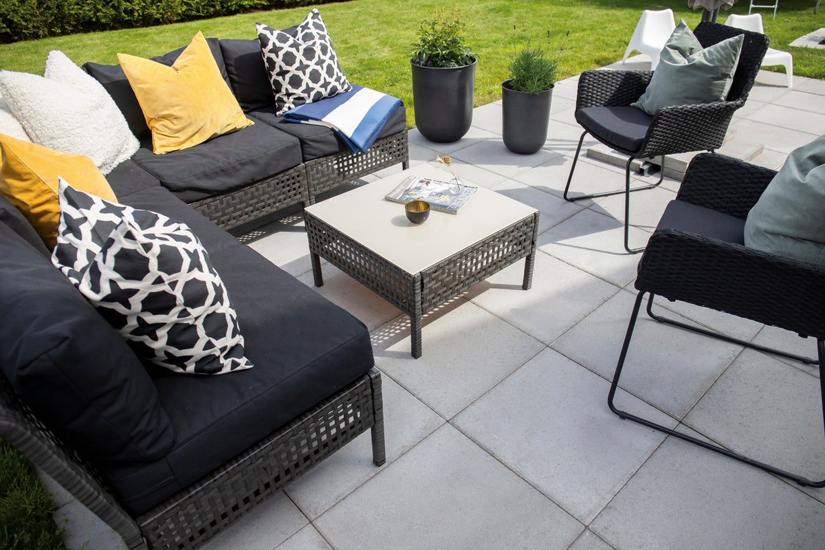 Loungeset bestående av hörnsoffa, två fåtöljer och ett kvadratiskt bord på en stenlagd terrass med kuddar och växter.