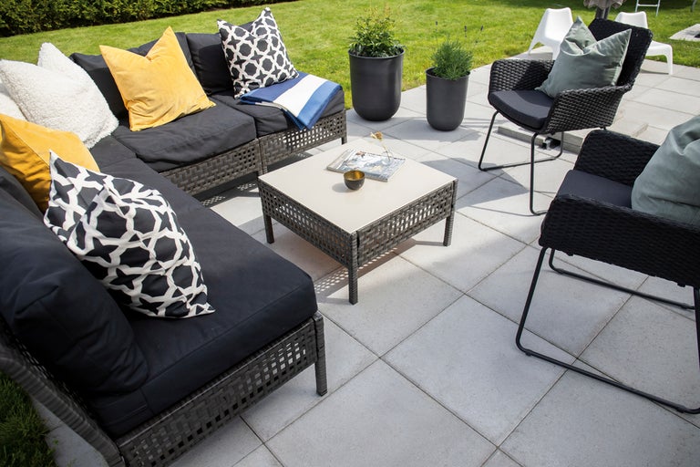 Loungeset bestående av hörnsoffa, två fåtöljer och ett kvadratiskt bord på en stenlagd terrass med kuddar och växter.