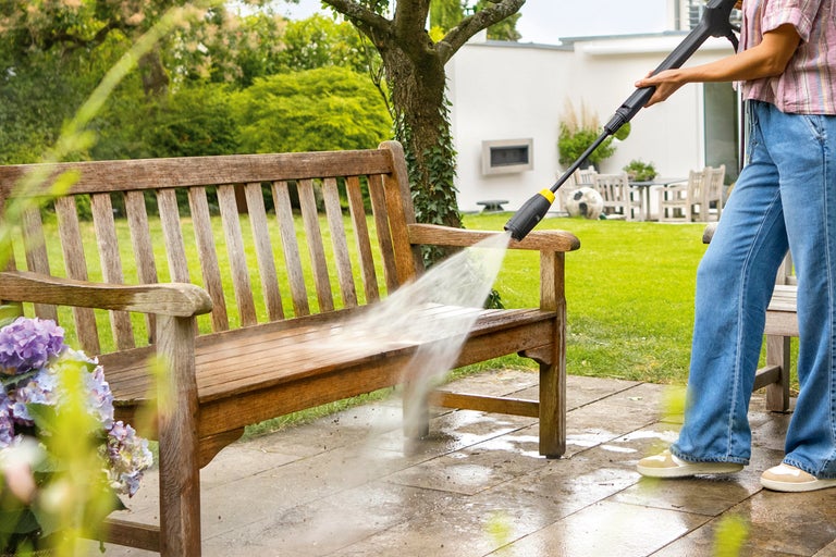 En person rengör en trädgårdsbänk i trä med en högtryckstvätt på en terrass.