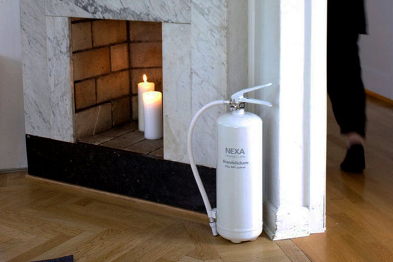 Interiörbild med en vit Nexa brandsläckare bredvid en öppen spis med brinnande ljus.