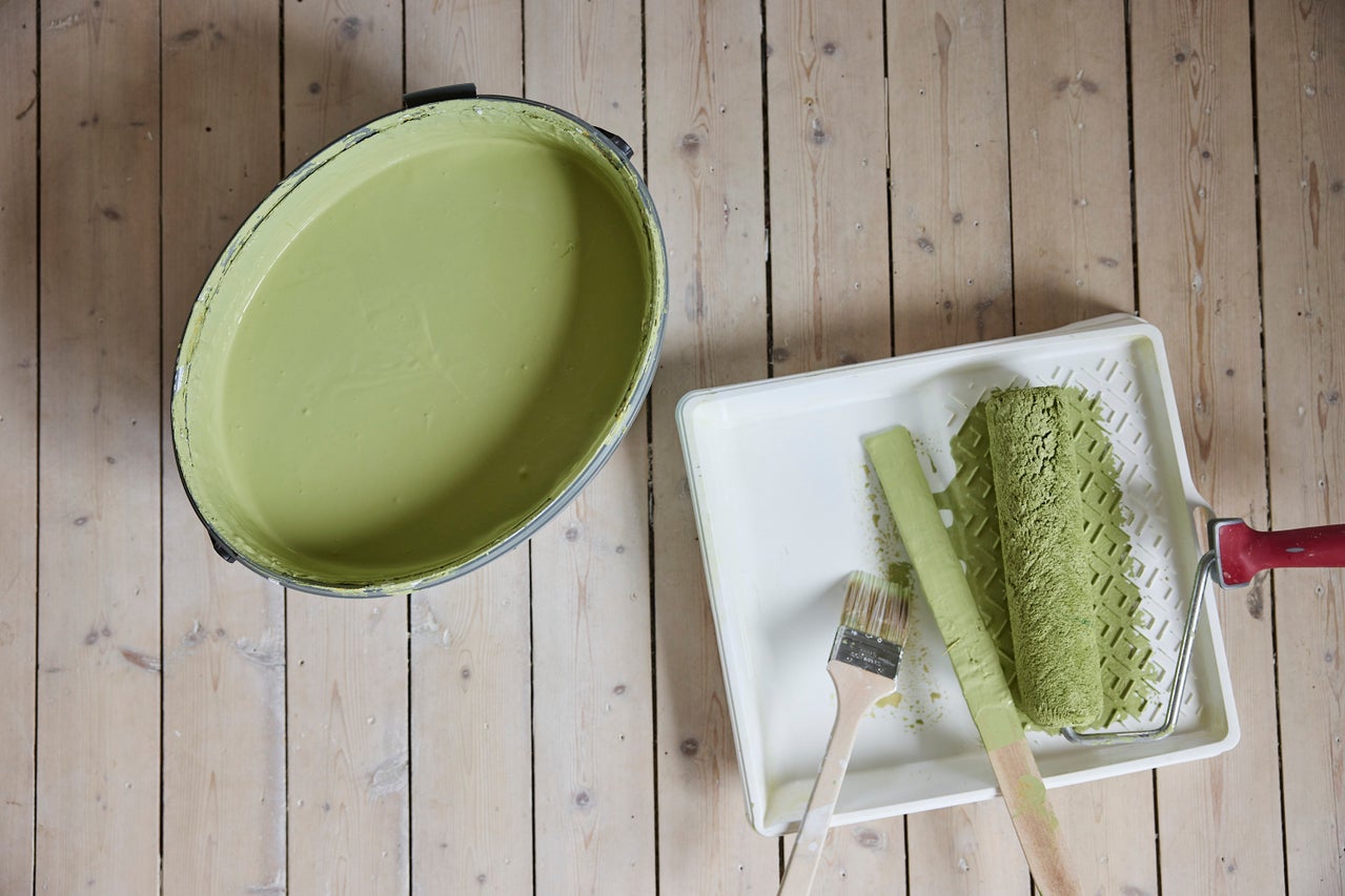 Hink med ljusgrön väggfärg, färgtråg med färgroller, pensel och rörsticka på trägolv.