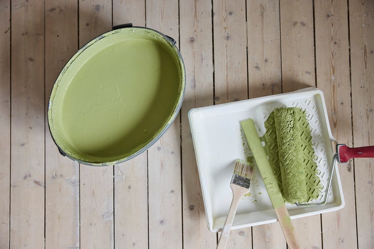 Hink med ljusgrön väggfärg, färgtråg med färgroller, pensel och rörsticka på trägolv.