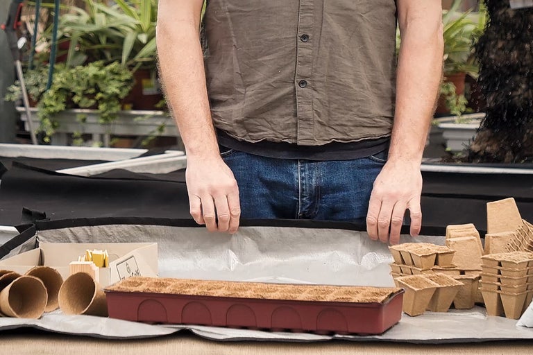 En person förbereder ett bord med såbrickor, krukor och planteringsjord för trädgårdsarbete.