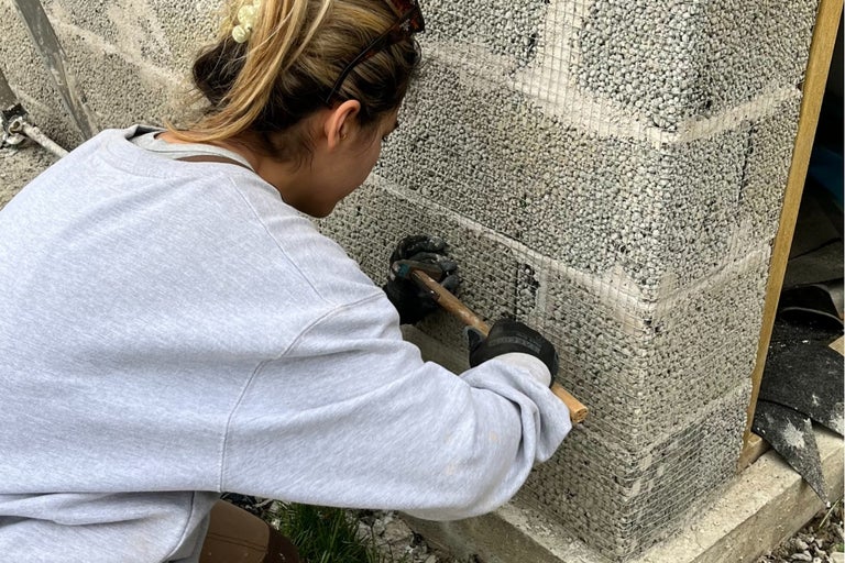 En person fäster ett trådnät på en mur av natursten med hjälp av en hammare.