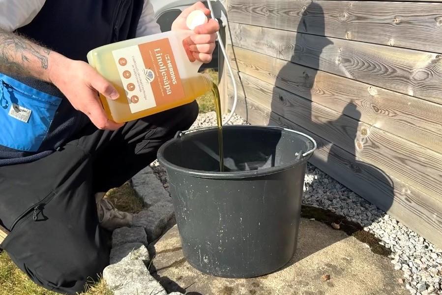 En person häller Herdins Linoljesåpa från en dunk i en hink bredvid en trävägg.