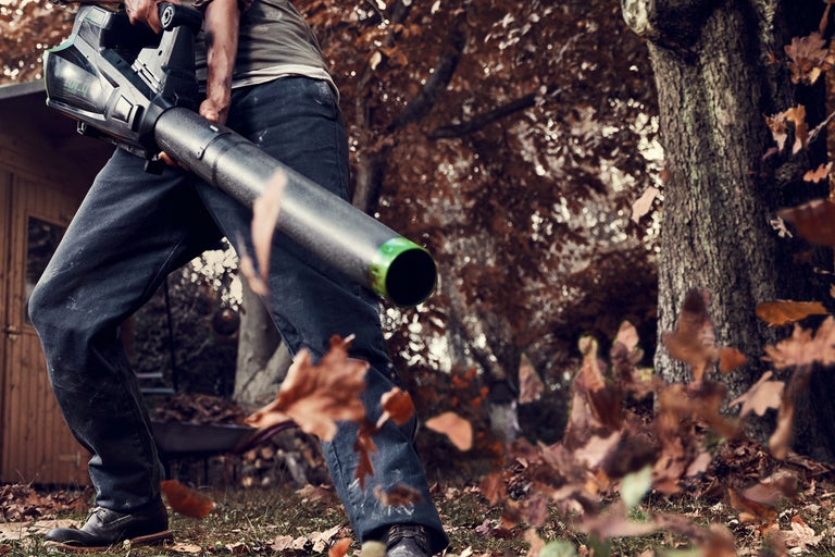 En person använder en lövblås i trädgården för att ta bort löv.