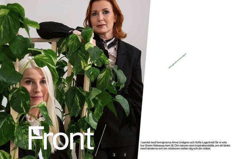 Två kvinnor poserar bakom en rumsavdelare i trä med gröna växter.