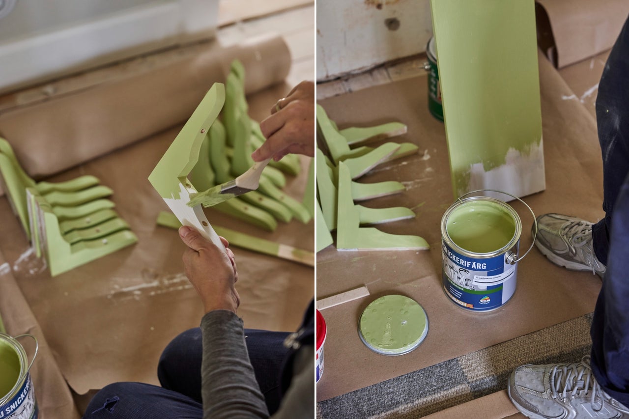 En person målar konsoler i trä med grön snickerifärg och en pensel bredvid en öppen färgburk.