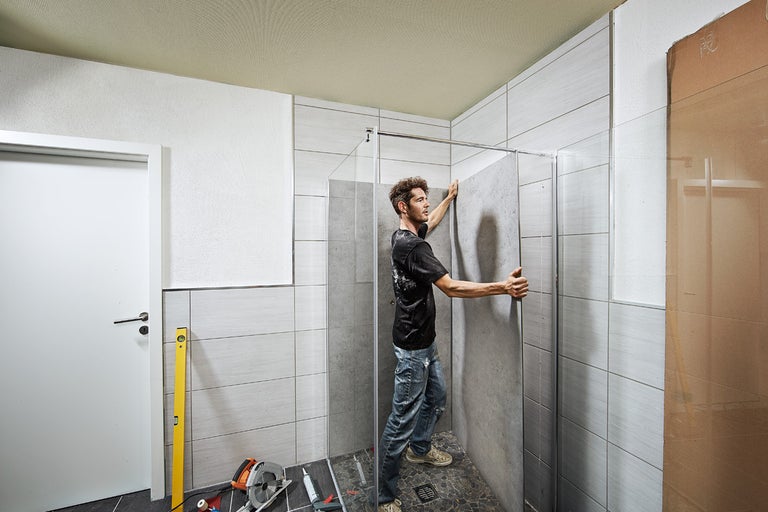 En man monterar en duschkabin i ett badrum med kakelväggar.