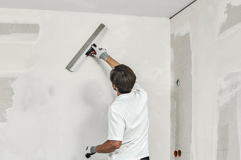 En man jämnar ut en vägg med en slätspackel i ett rum.