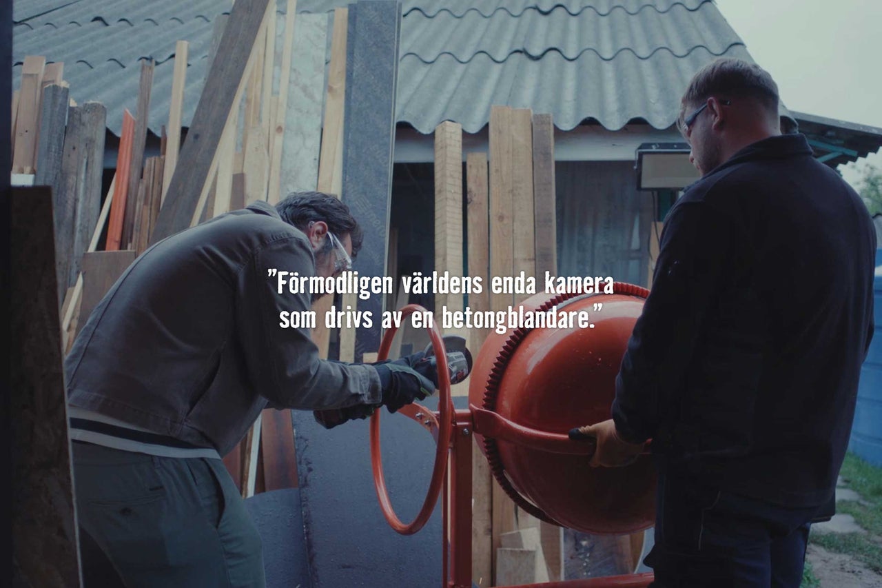 Två hantverkare arbetar med en betongblandare i en verkstadsmiljö med lagrade träplankor.