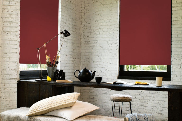 Scen med två fönster med röda rullgardiner, bord med föremål och sittplatser i ett ljust rum med tegelväggar