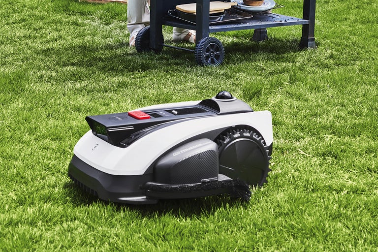 Modern robotgräsklippare i vitt och svart på en gräsmatta bredvid en grillvagn i trädgården.