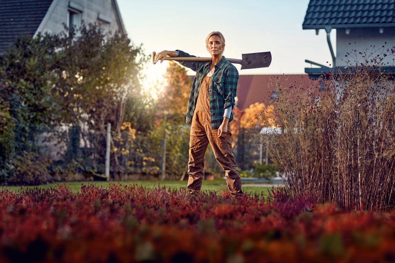 Kvinna i trädgården med spade på axeln framför huset