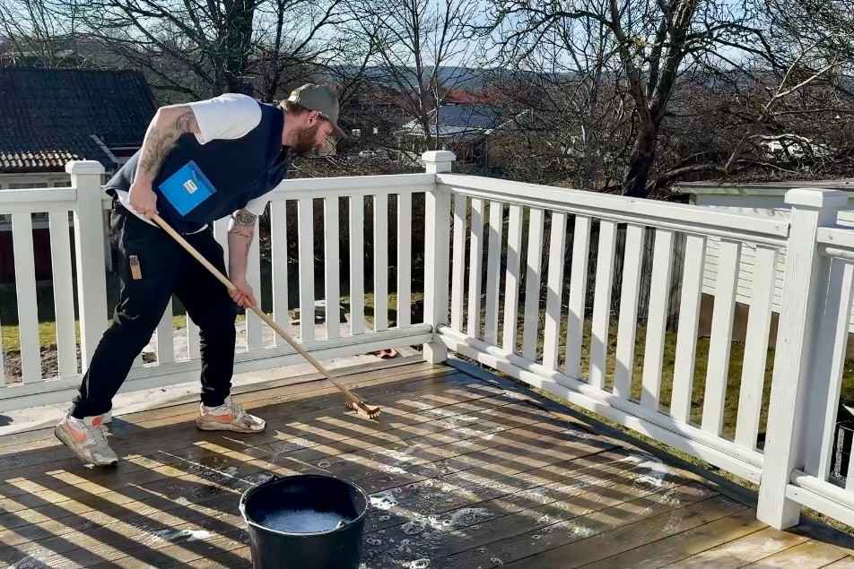 En man rengör en träterrass med en skurborste och såpvatten. En hink står i förgrunden, omgiven av ett vitt räcke.