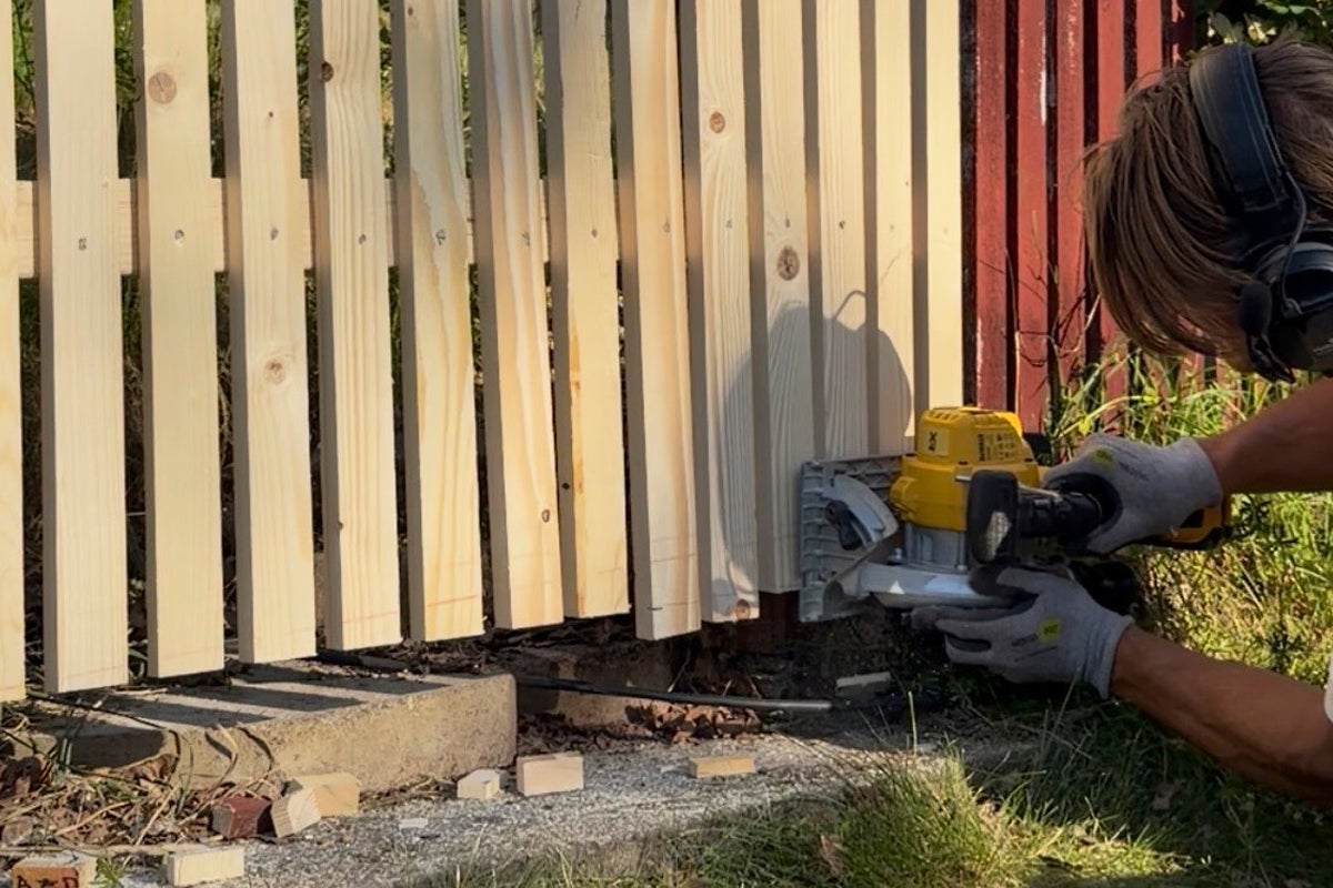 En person kapar spjälorna på ett ljust trästaket med en handcirkelsåg utomhus.