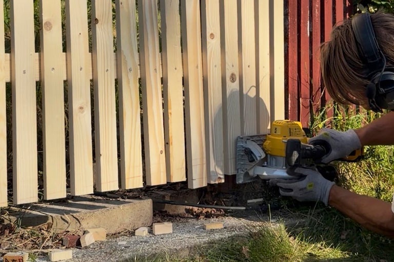 En person kapar spjälorna på ett ljust trästaket med en handcirkelsåg utomhus.
