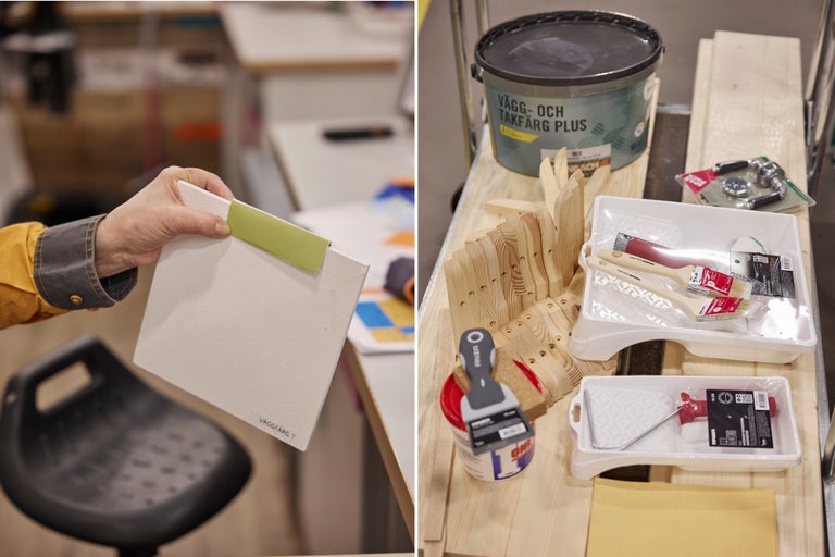 En hand håller ett vitt färgprov bredvid måleritillbehör som en färgburk, penslar, tråg och träkonsoler på en arbetsbänk.