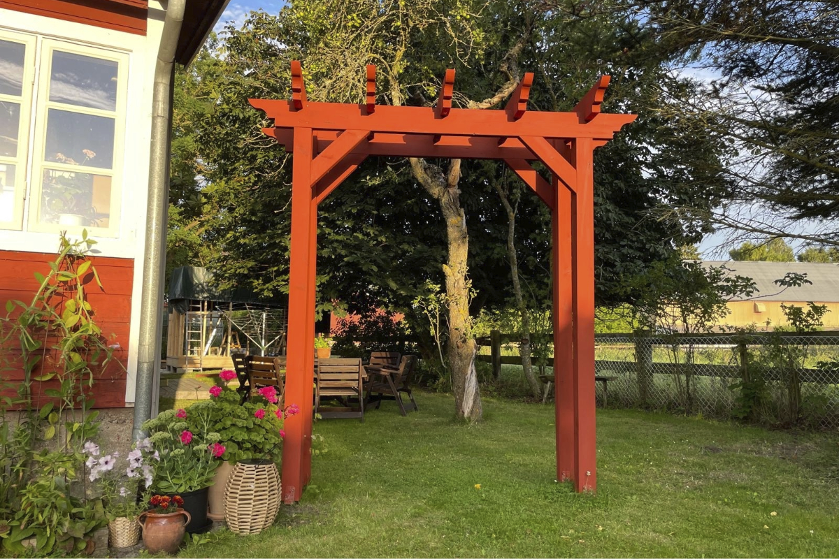 En röd träpergola som trädgårdsbåge står på en gräsmatta framför ett hus med trädgårdsmöbler i bakgrunden.